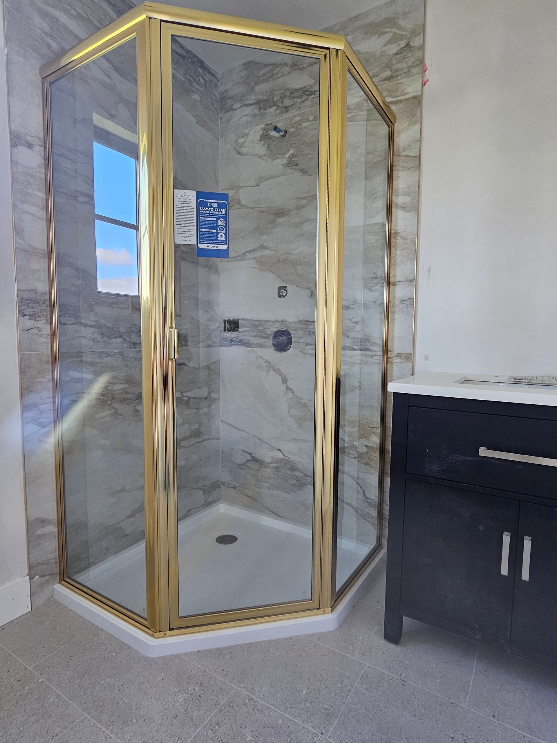 Gold-framed corner shower in a bathroom with gray tile walls and a dark vanity.