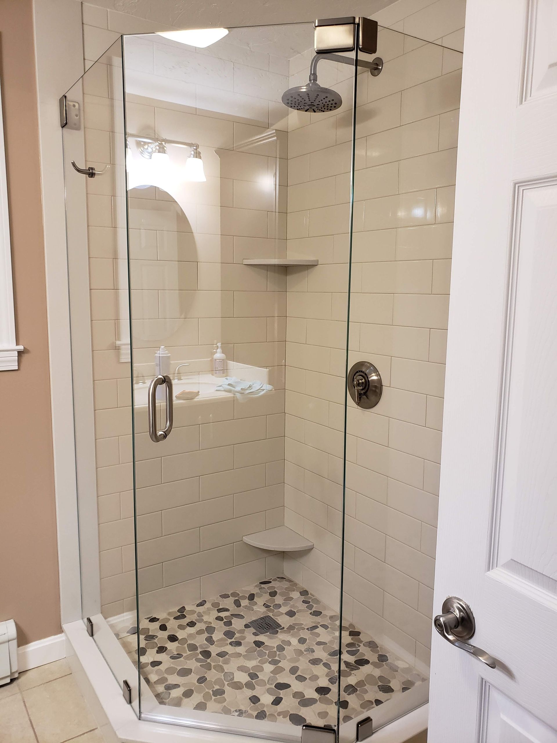 Glass-enclosed shower with pebble floor, beige tile walls, and a built-in shelf. A white door is on the right.