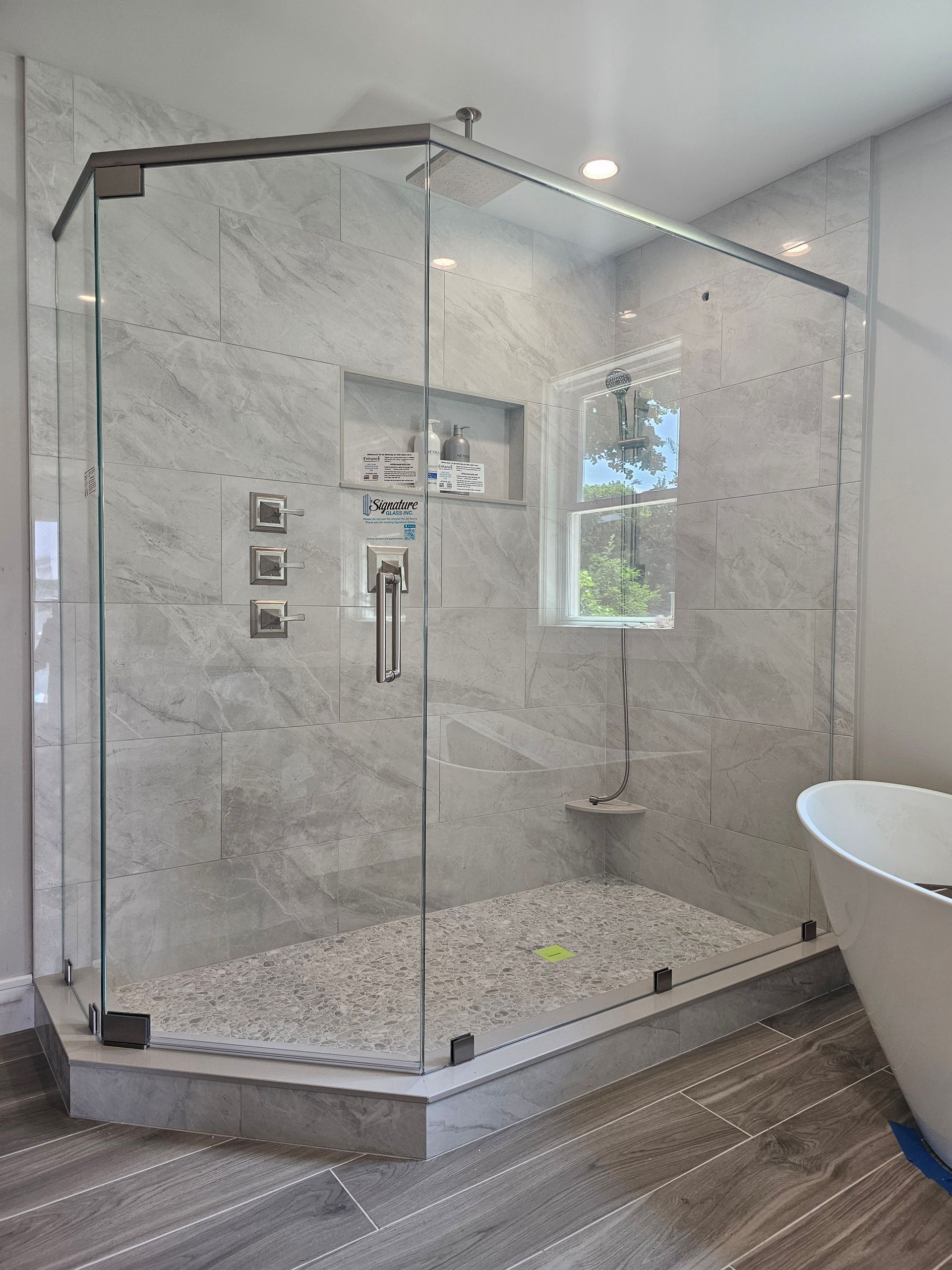 Glass shower with pebble floor, gray tiled walls, chrome fixtures, and a small window.