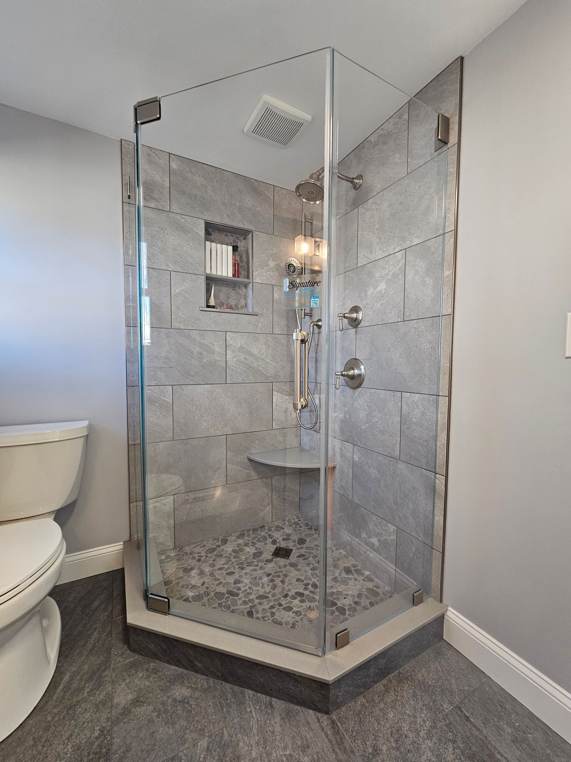 Corner shower with gray tile, glass doors, and pebble floor. Toilet and gray walls are also visible.