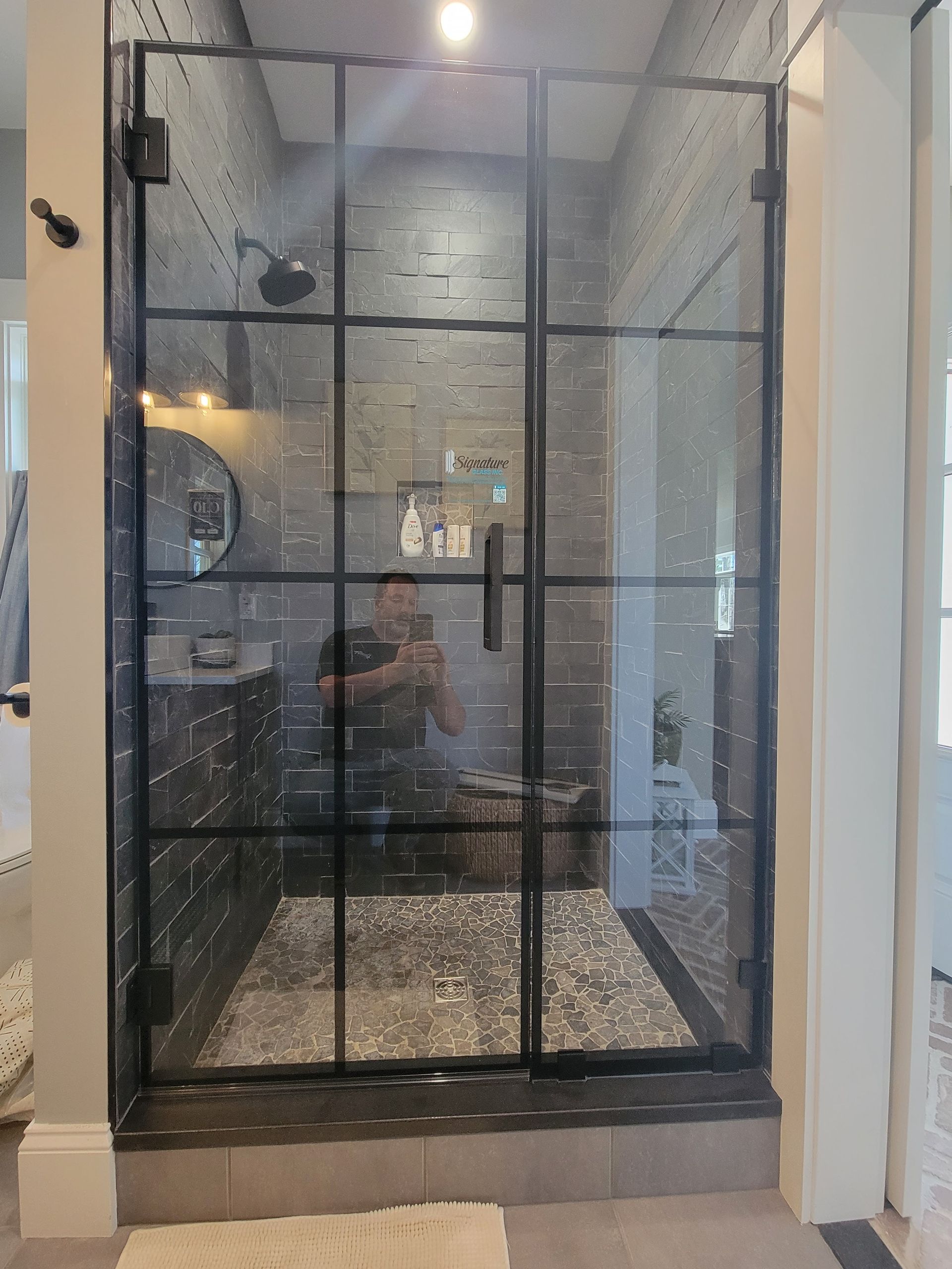 Black-framed glass shower door in a modern bathroom. Gray stone tile interior. Someone reflected in the glass.