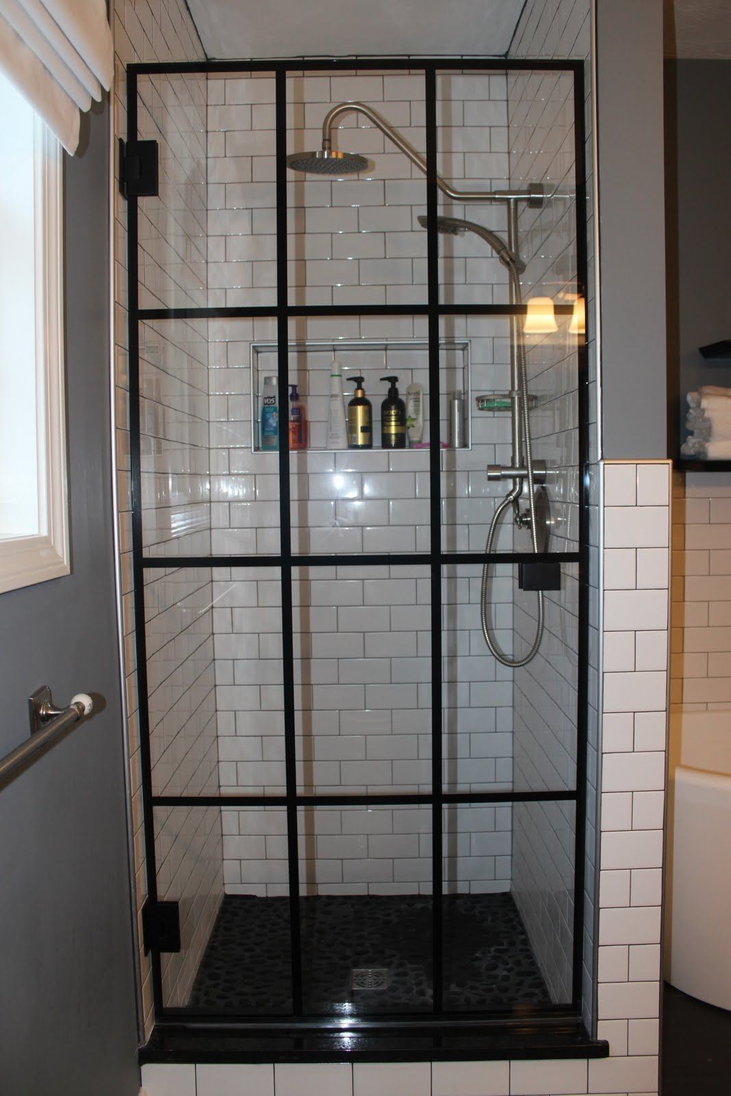 Black-framed shower with grid design. White subway tiles, black floor, and a built-in shelf.