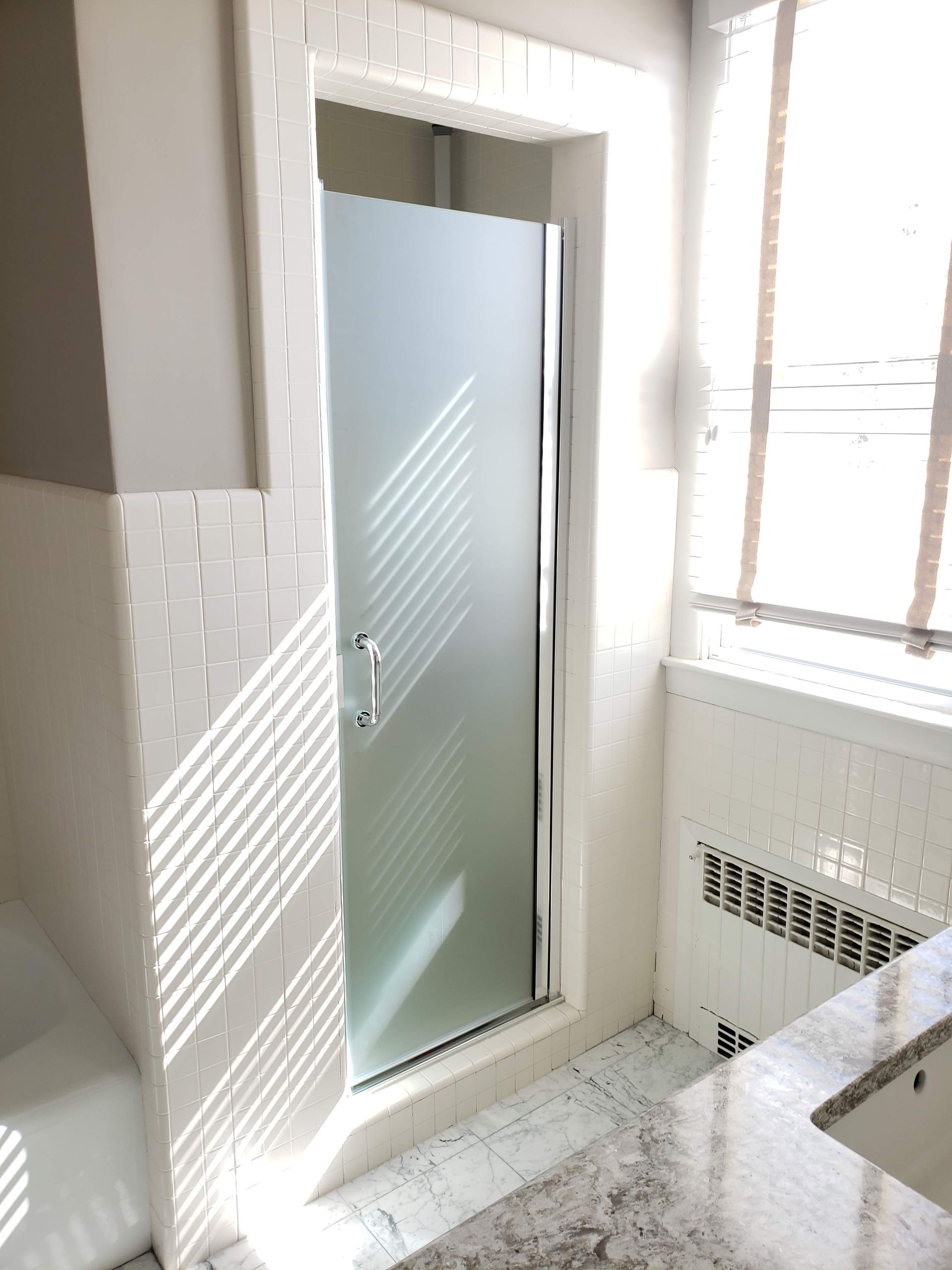 Shower with frosted glass door in a bathroom with white tile and a window with blinds.