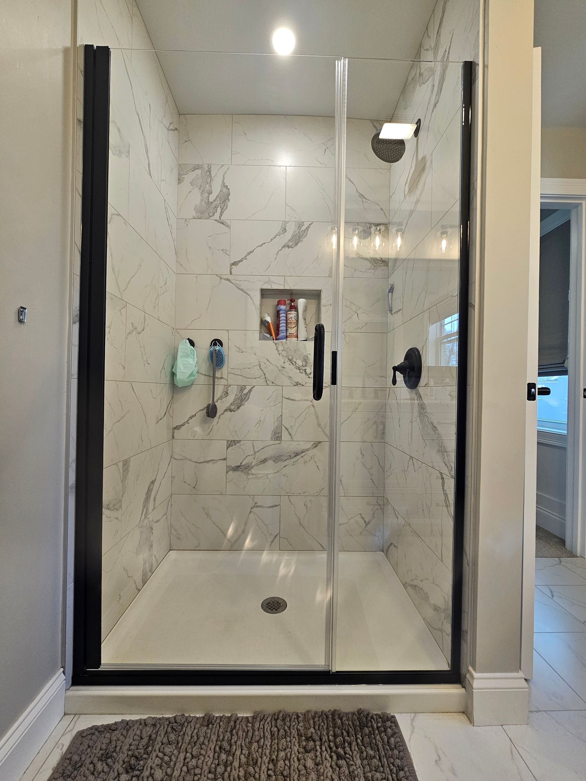 Shower with white and gray marble tile, black frame, glass door, toiletries, and brown rug.