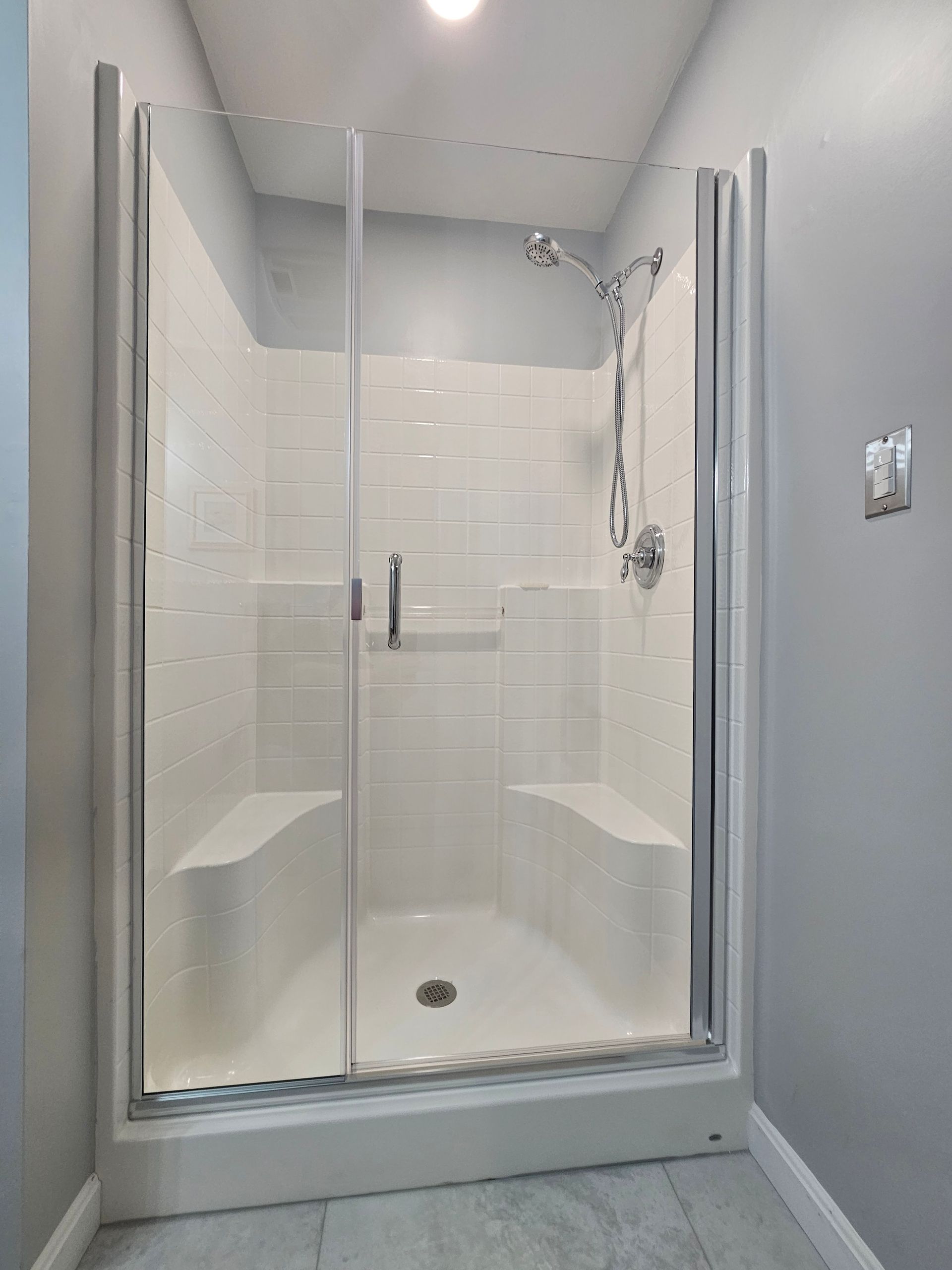 Shower with glass doors, white tiled walls, two built-in benches, and silver fixtures in a bathroom.
