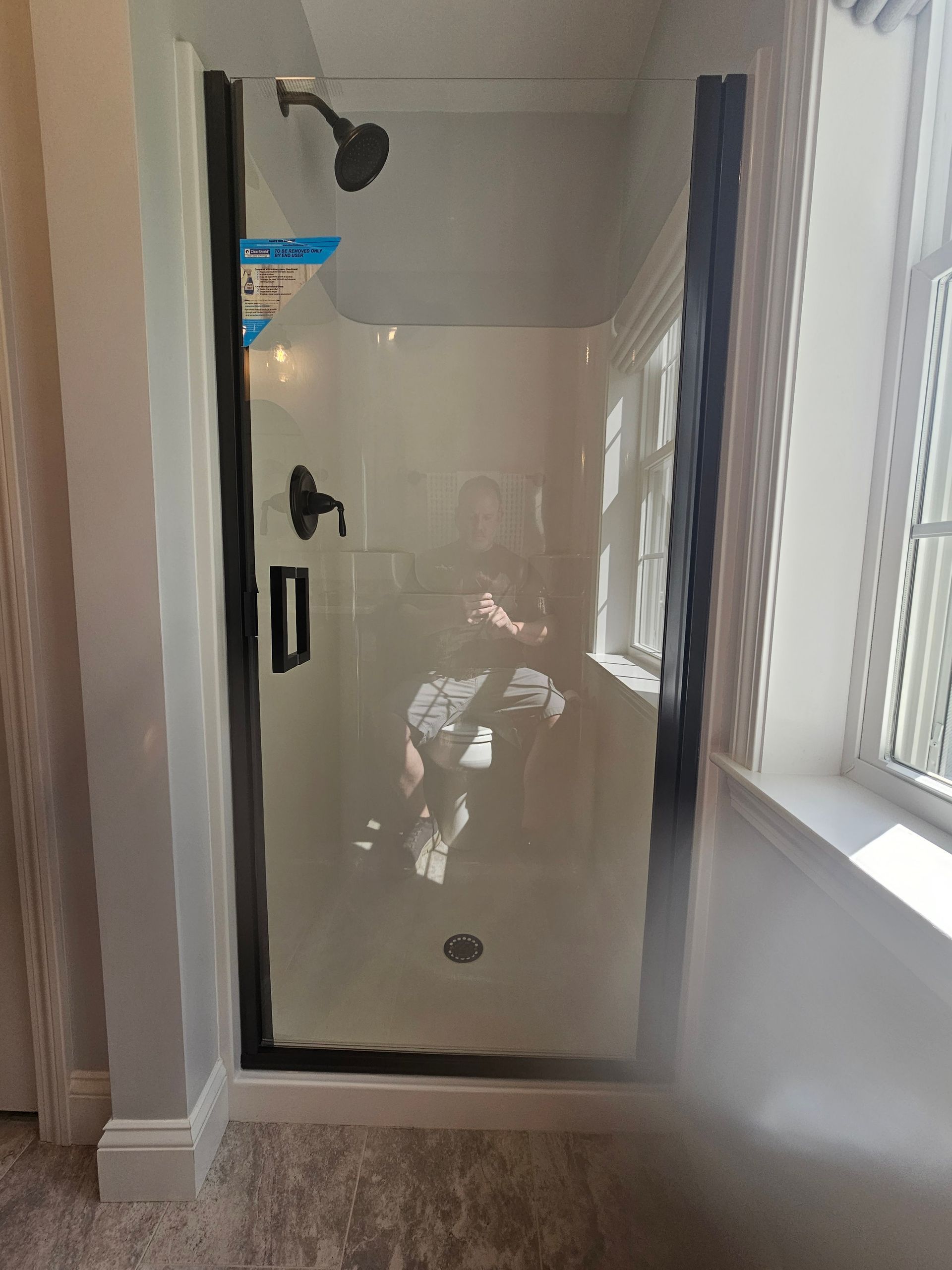 Black-framed glass shower enclosure with black fixtures. Window on the right. Person reflected in the glass.