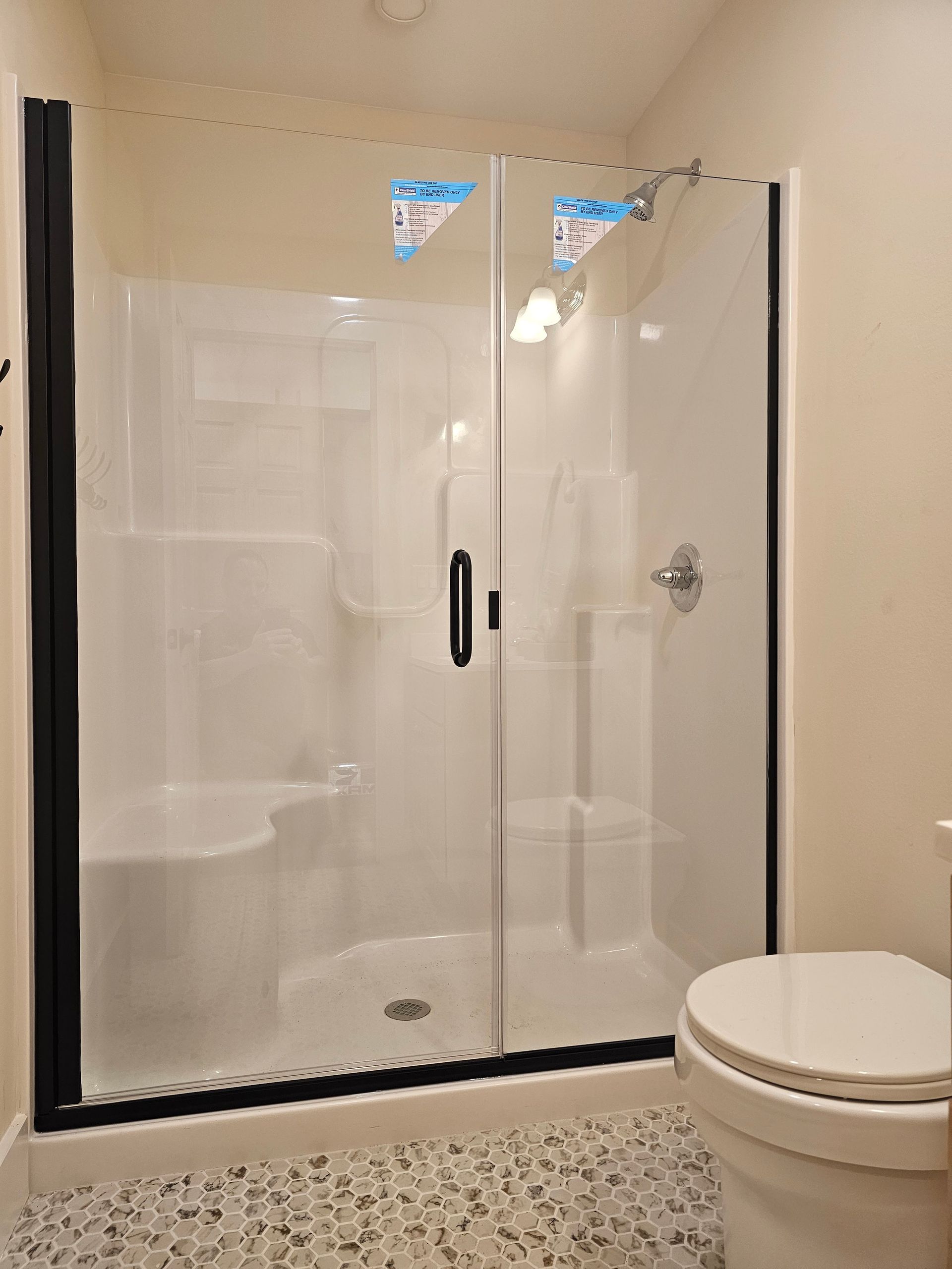 White shower with black trim and sliding glass doors, next to a toilet, on a speckled tile floor.