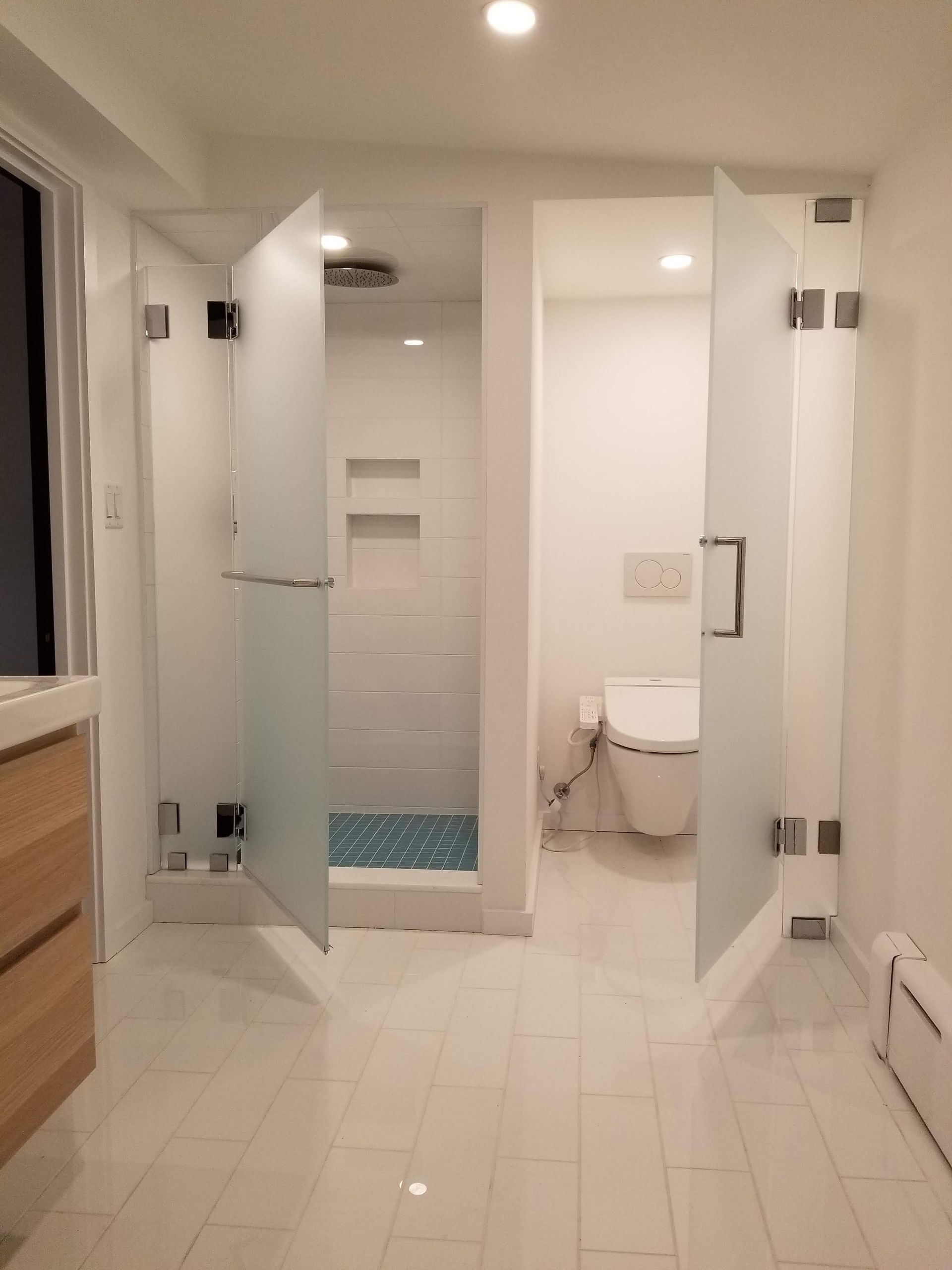 Modern bathroom with frosted glass shower and toilet stalls, white tile floors, and light-colored wood vanity.