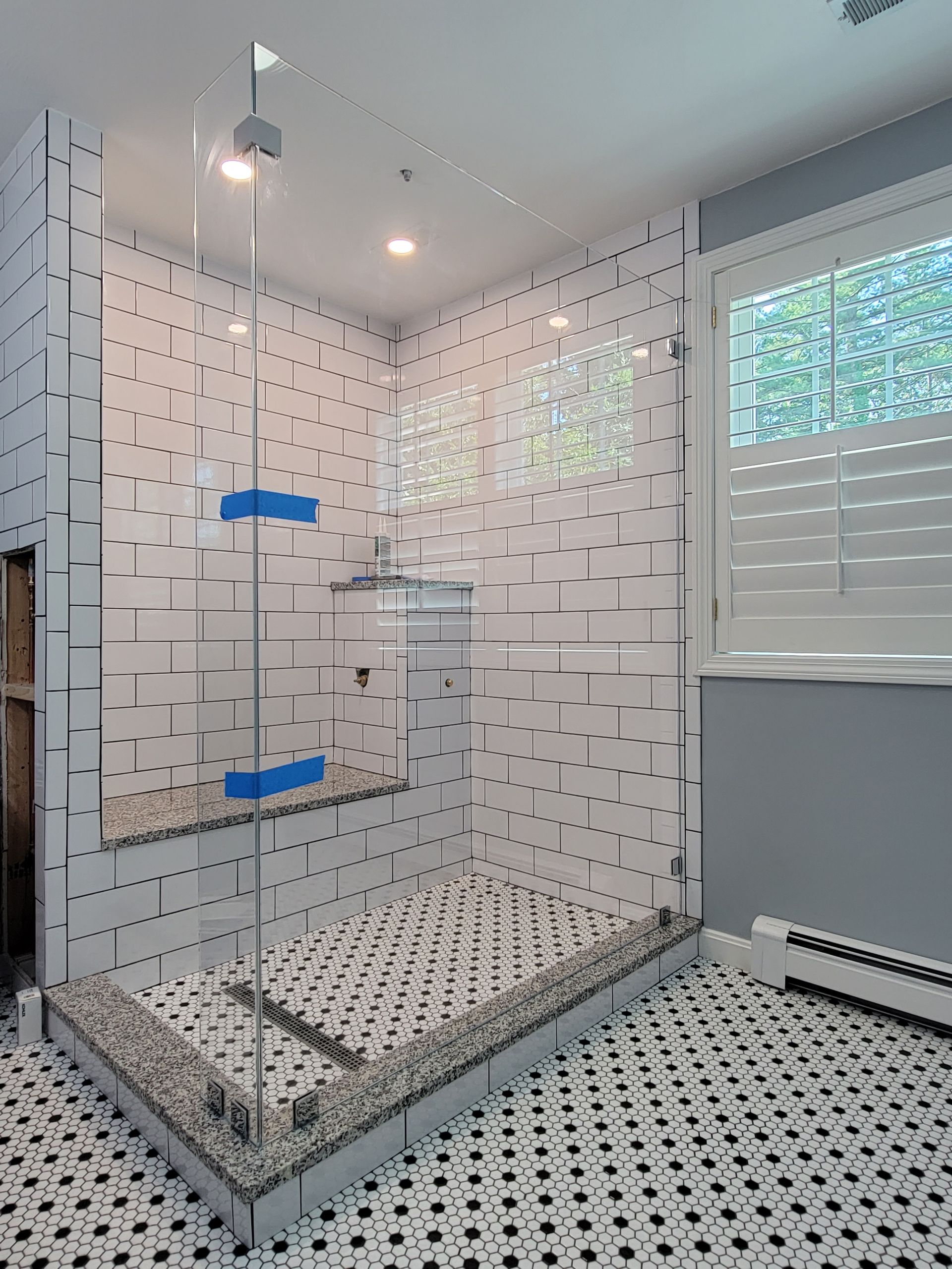 Corner shower with glass doors, white brick-like tiles, pebble shower floor, black and white tiled bathroom floor, window.