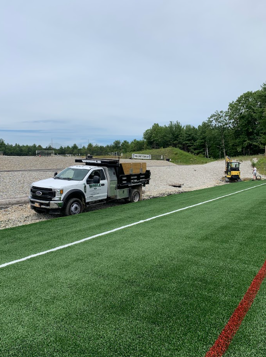 Two utility trucks on a turf field beside a gravel berm under a cloudy sky