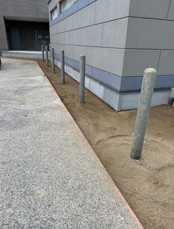 Row of metal bollards along a building edge beside a concrete walkway and dirt strip