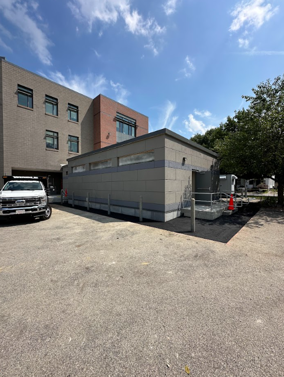 Small gray portable building beside an apartment, with a white truck and orange cones in a gravel lot under a blue sky