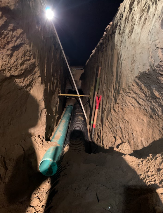 Narrow trench at night with a large green pipe and tools between tall dirt walls under a bright light