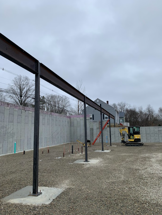 Gravel lot with steel posts and an overhead beam under construction beside a concrete wall, with a yellow excavator.