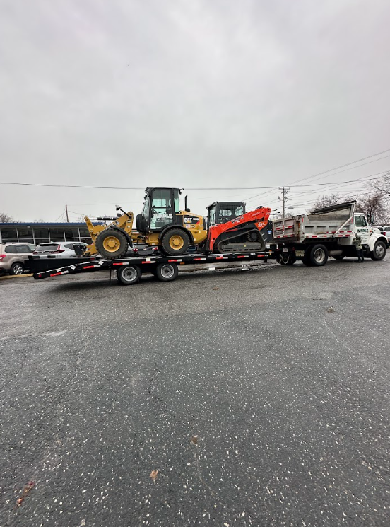 Flatbed tow truck carrying a yellow loader and orange excavator on a wet road under a gray sky