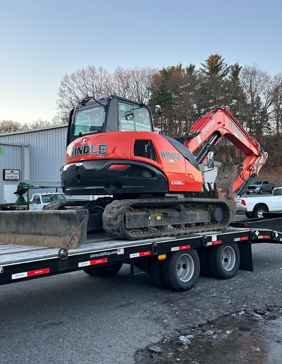 Orange excavator on a flatbed trailer in a parking lot, with trees and vehicles in the background.