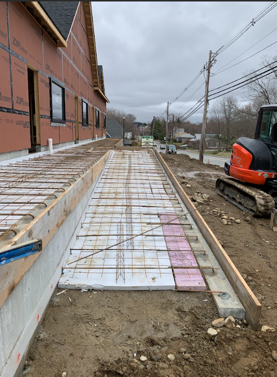 Construction site beside brick building with a narrow poured walkway and excavator on the right