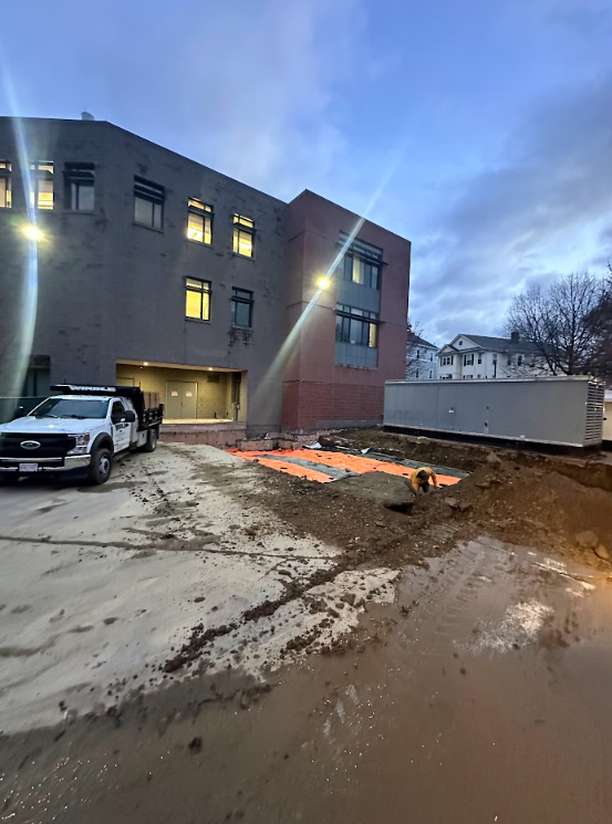 Gray building under construction at dusk, with muddy ground, a white truck, and an orange-lit work area.