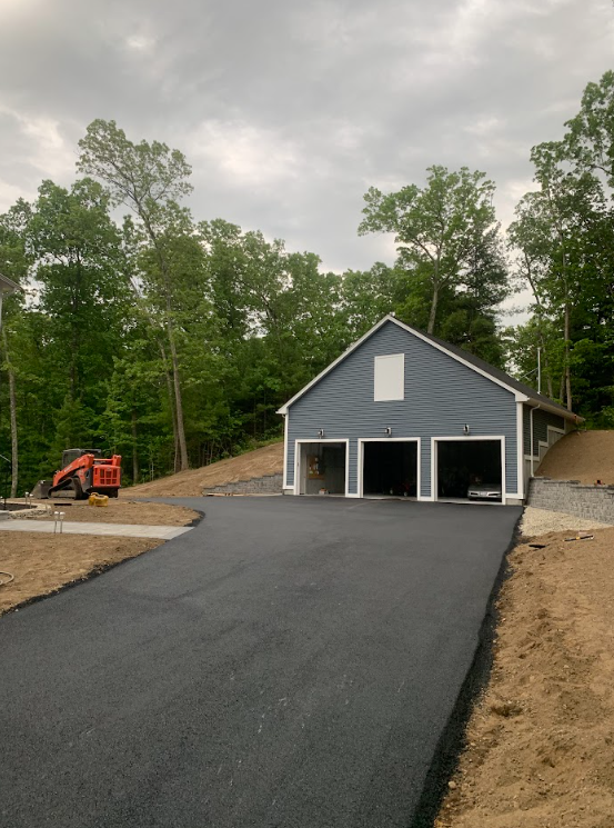 Newly paved driveway leading to a gray garage under construction, with a small excavator nearby and trees behind.