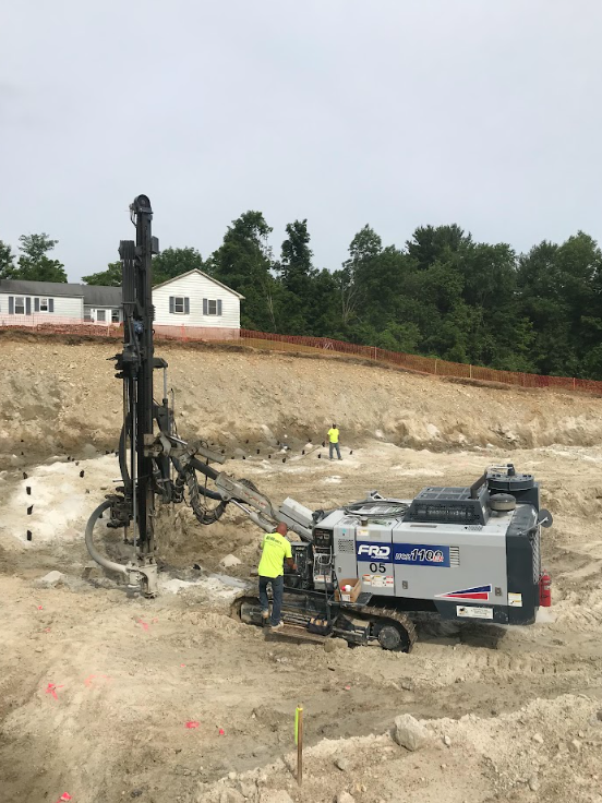 Construction crew drilling rock on a hillside, with heavy equipment and a white truck near a house above.