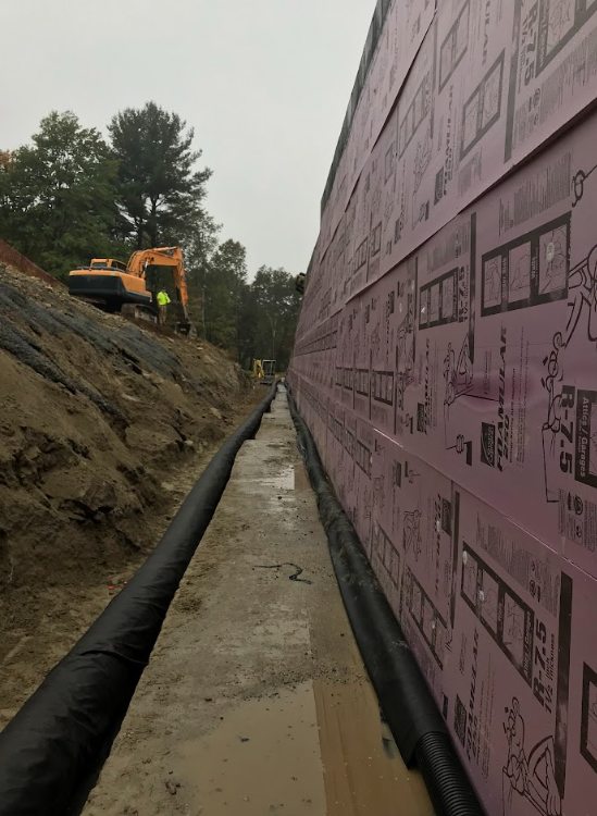 Construction trench beside pink insulated wall, with an excavator working on the left.