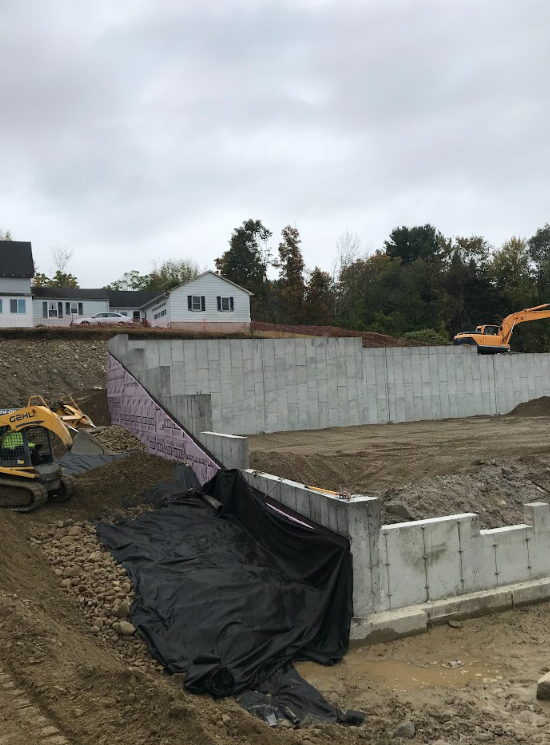 Construction site with concrete retaining wall, black tarp, and excavator under overcast sky