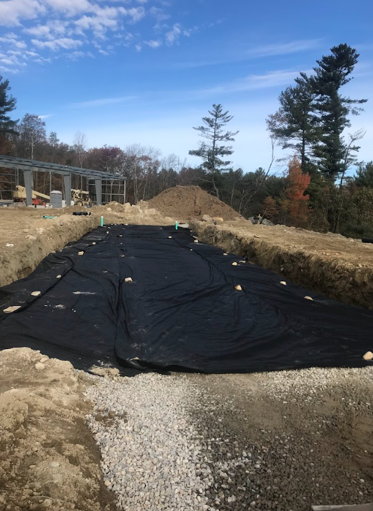 Construction site with black silt barrier over gravel trench, dirt mounds, and trees under a blue sky