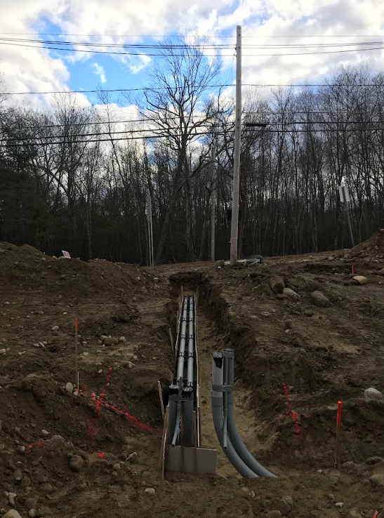 Utility conduits emerging from a dirt trench at a wooded construction site under a cloudy sky