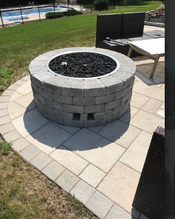 Stone fire pit on a paved patio beside a lawn, with pool and seating nearby.
