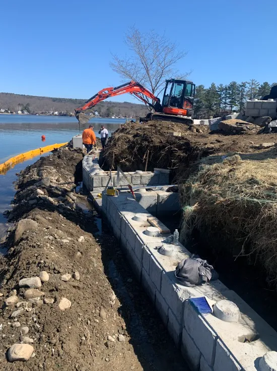 Excavator and workers reinforcing a shoreline retaining wall beside water under a clear blue sky