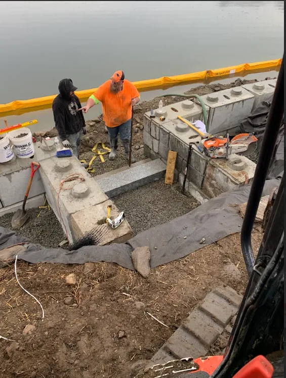 Two workers in rain gear stand in a concrete foundation trench beside a yellow barrier