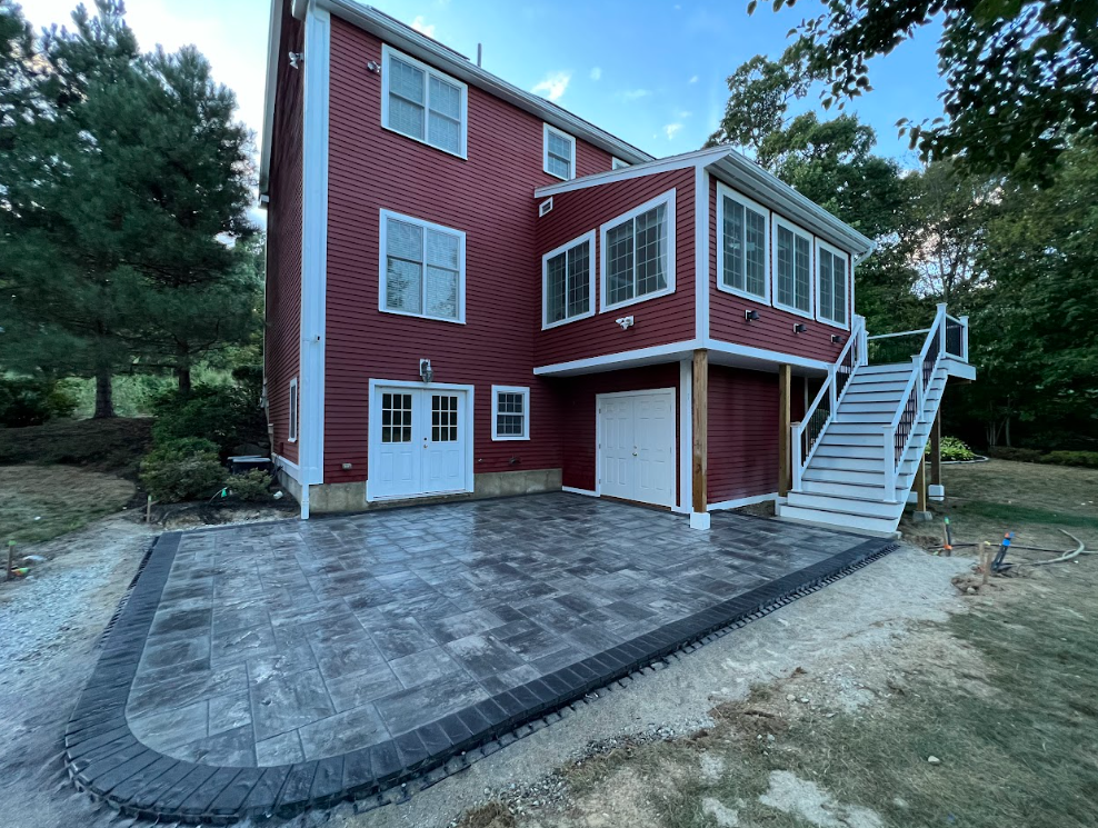 Red two-story house with large paved patio and exterior stairs in a wooded yard
