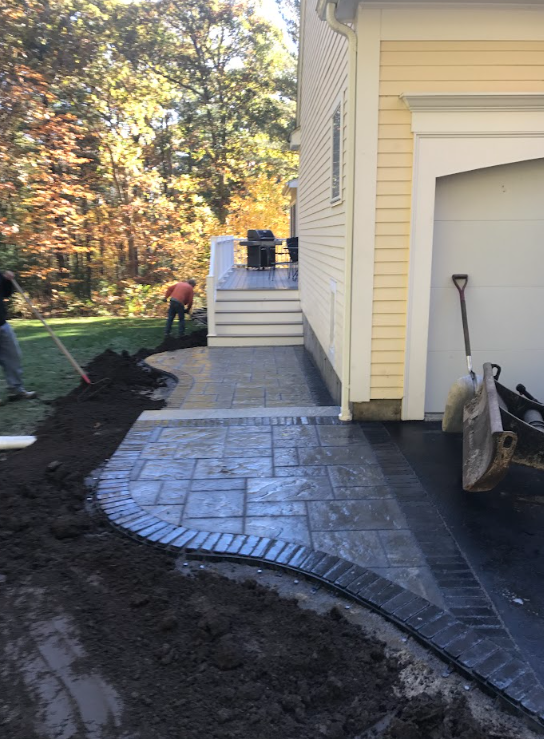 Partially completed patio beside a yellow house, with workers and landscaping soil nearby