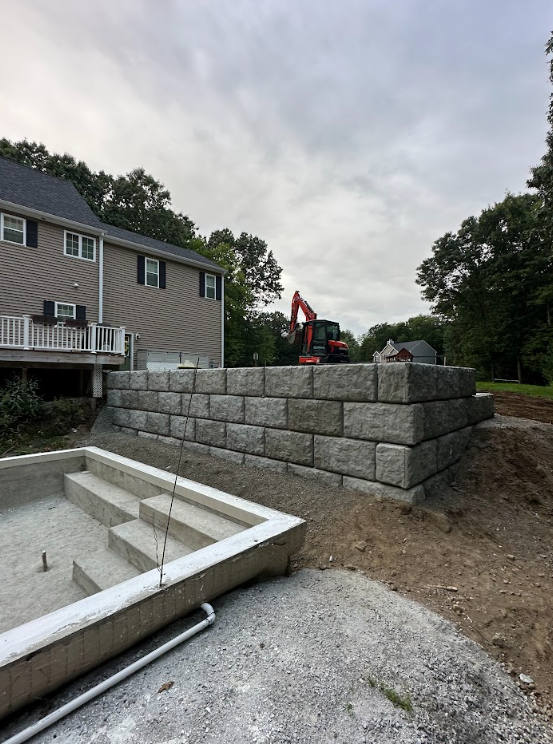 Construction site with stacked retaining wall blocks beside a house and an excavator in the background
