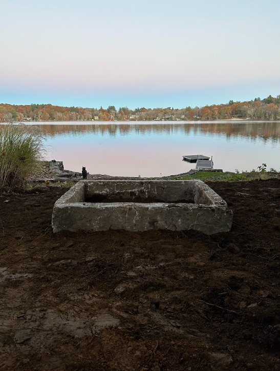 Stone fire pit on a lakeshore at dawn, with calm water, a small dock, and a tree-lined horizon.