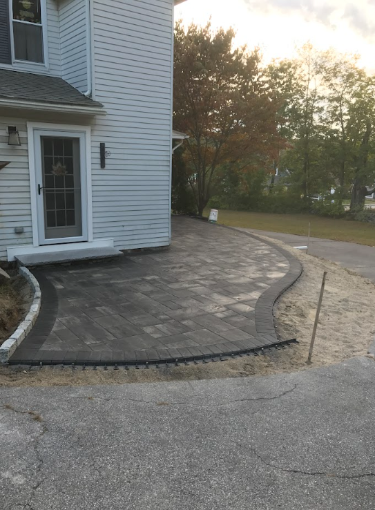Curved brick patio beside a white house, with a driveway and trees at sunset.