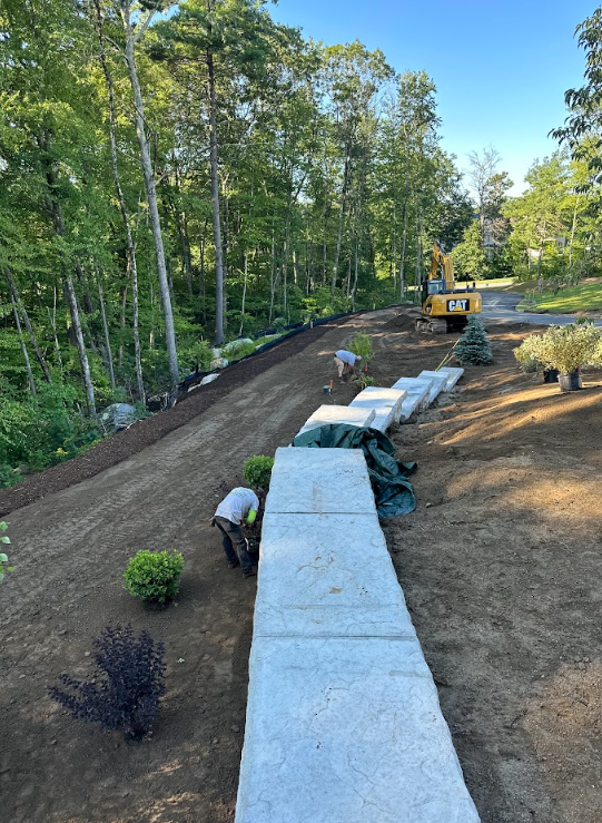 Workers laying a long white retaining wall beside a dirt slope in a wooded yard