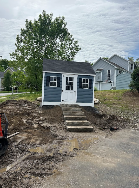 Small blue shed with white trim on a dirt lot, with steps leading up to the door and a tree behind it
