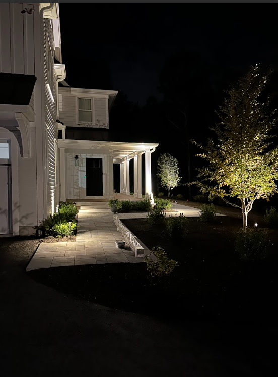 Nighttime view of a house exterior with a lit porch, walkway, and illuminated tree in the yard
