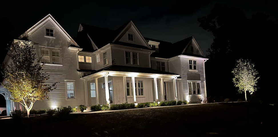 Large white house at night, warmly lit with exterior lights and glowing windows.