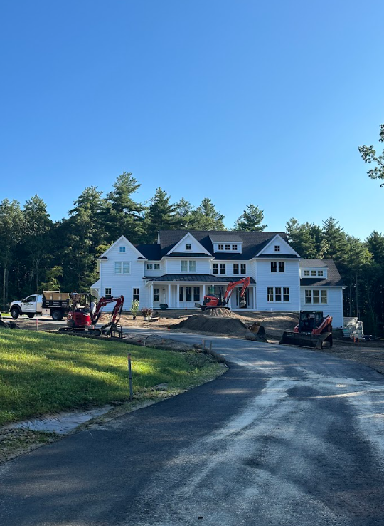 Large white house under construction with scaffolding, tools, and vehicles on a curved driveway under a blue sky