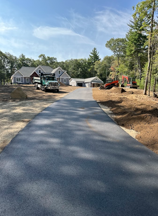 Freshly paved driveway leading to a house under construction with trees and construction equipment nearby
