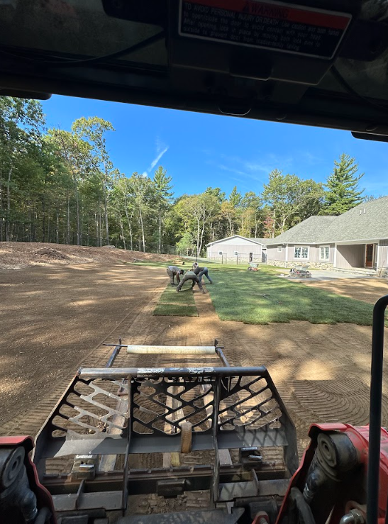 View from a utility vehicle over a dirt lot toward a white house and trees under a blue sky