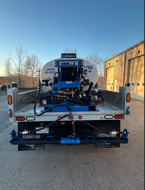 Blue industrial machine mounted on a flatbed trailer, parked outdoors beside a building.