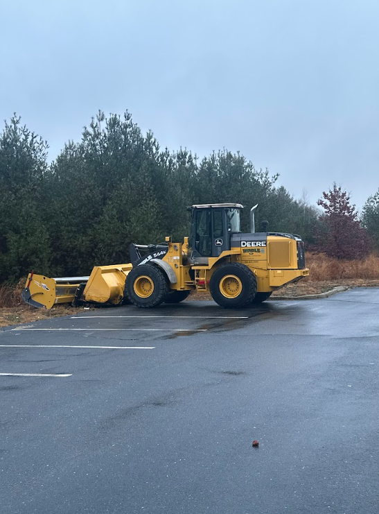 Yellow front loader parked on wet pavement beside evergreens under a cloudy sky