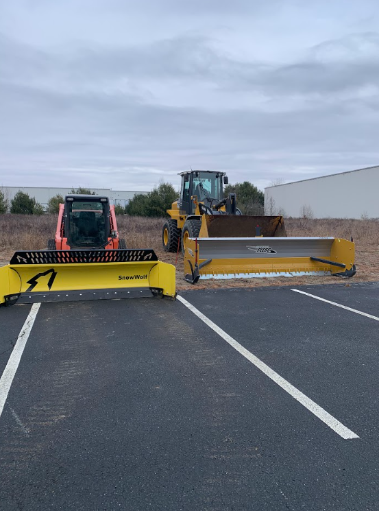 Two snowplows parked in a lot, one orange and black, one yellow, under a cloudy sky