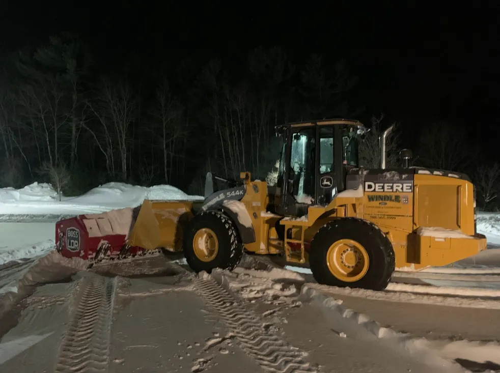 Yellow John Deere loader pushing snow at night in a snowy field beside a red container