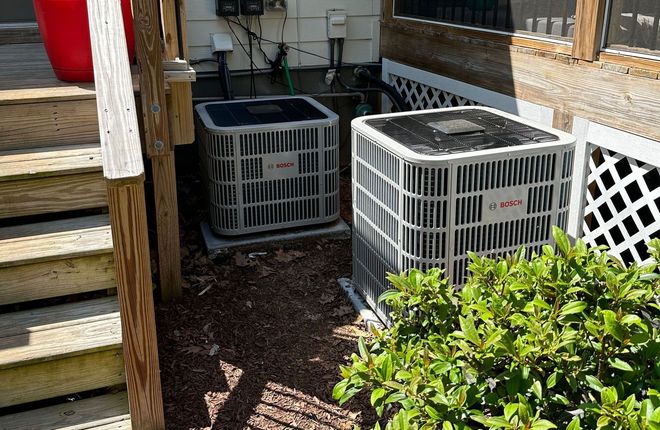 Two air conditioning units near a house. They are gray, with a brown mulch bed.