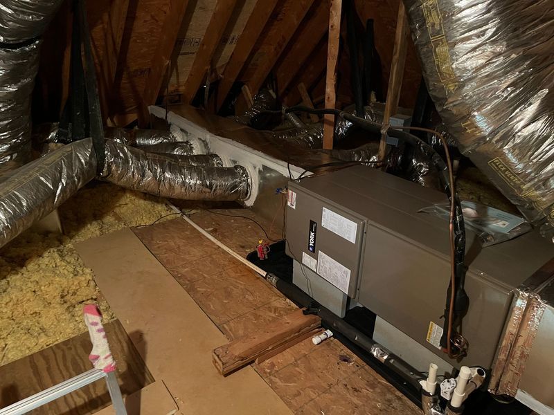 HVAC unit in an attic with ductwork, insulation, and wooden planks.