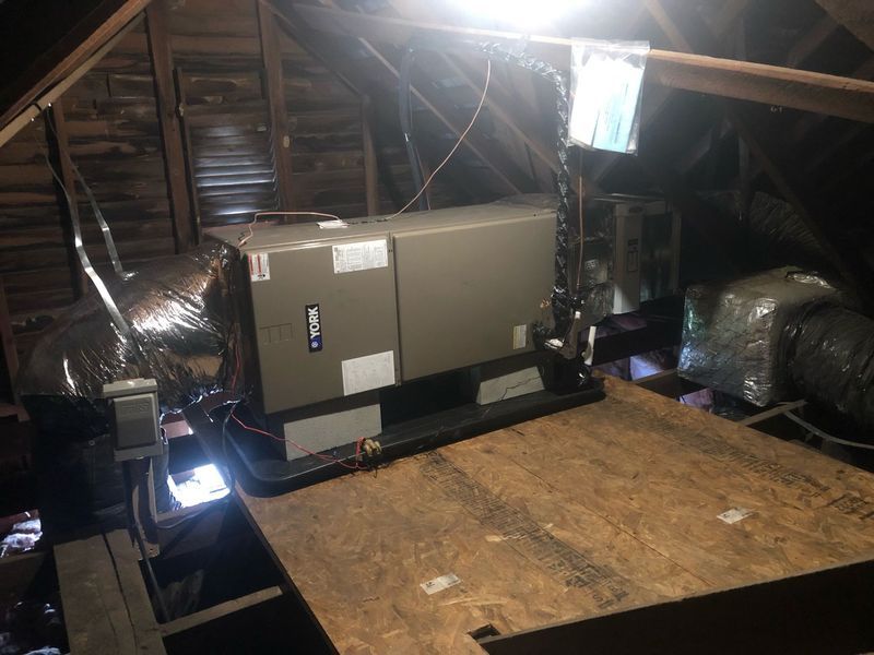 HVAC unit in an attic. Ductwork, electrical wiring, and a plywood base are visible. Dim lighting.